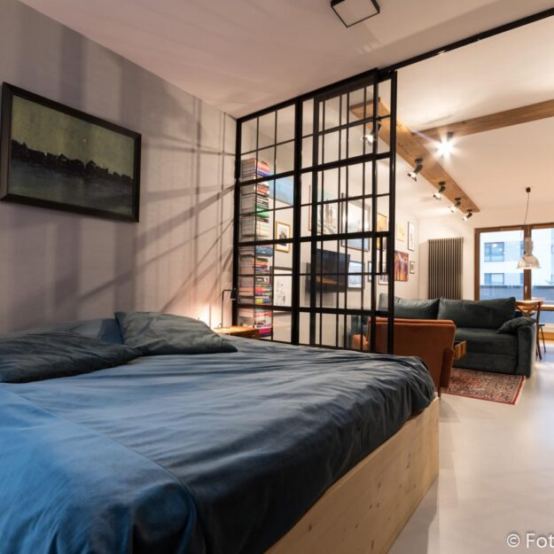 Zdjęcie aranżacji wnętrz sypialni w mieszkaniu zaprojektowanym w stylu industrialnym. Sypialnia połączona z salonem za pomocą ażurowej przeszklonej ścianki z czarnymi szprosami.