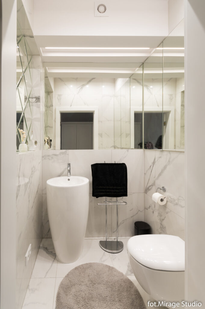Zdjęcie aranżacji małej toalety w stylu glamour w jasnych kremowo białych kolorach. Ściany wykończone płytkami imitującymi biały marmur, który przeplata się z obszernymi lustrami optycznie powiększającymi WC