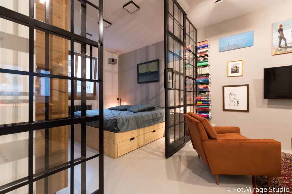 industrialne-wnetrze-mieszkanie-bielany-11 Styl loftowy, industrialna sypialnia w mieszkaniu na Bielanach – drewniane łóżko z szufladami, przeszklenia loftowe, ściana z książkami i pomarańczowy fotel