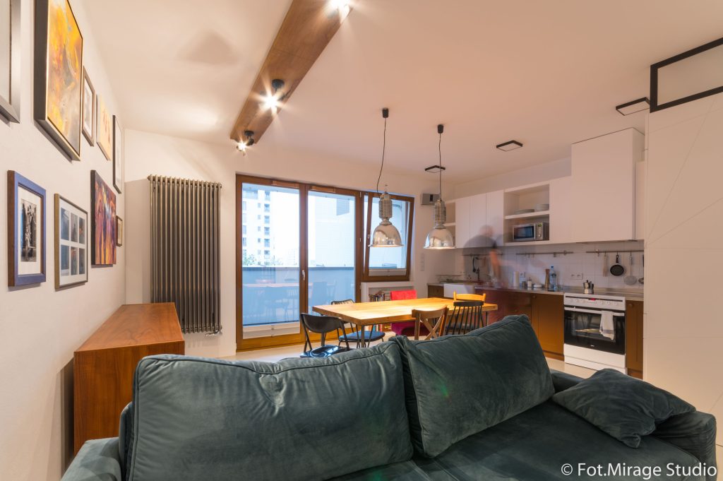 Industrialne wnętrze mieszkania na Bielanach – salon z kuchnią, drewniane belki, metalowe lampy i zielona sofa